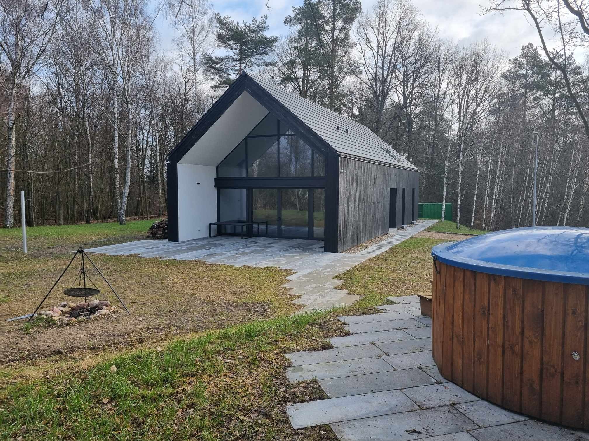 Nowoczesny dom z ciemnego drewna z dużymi przeszkleniami, otoczony lasem, z wybrukowanym tarasem i jacuzzi na zewnątrz.