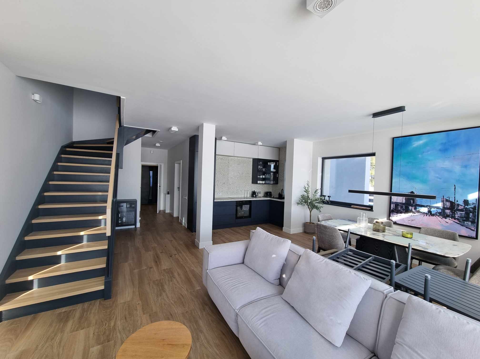 Jasne, przestronne wnętrze domu w Komorowie: drewniane schody z ciemnymi podstopnicami, parkiet, szara sofa i otwarta kuchnia z ciemnymi szafkami dolnymi.