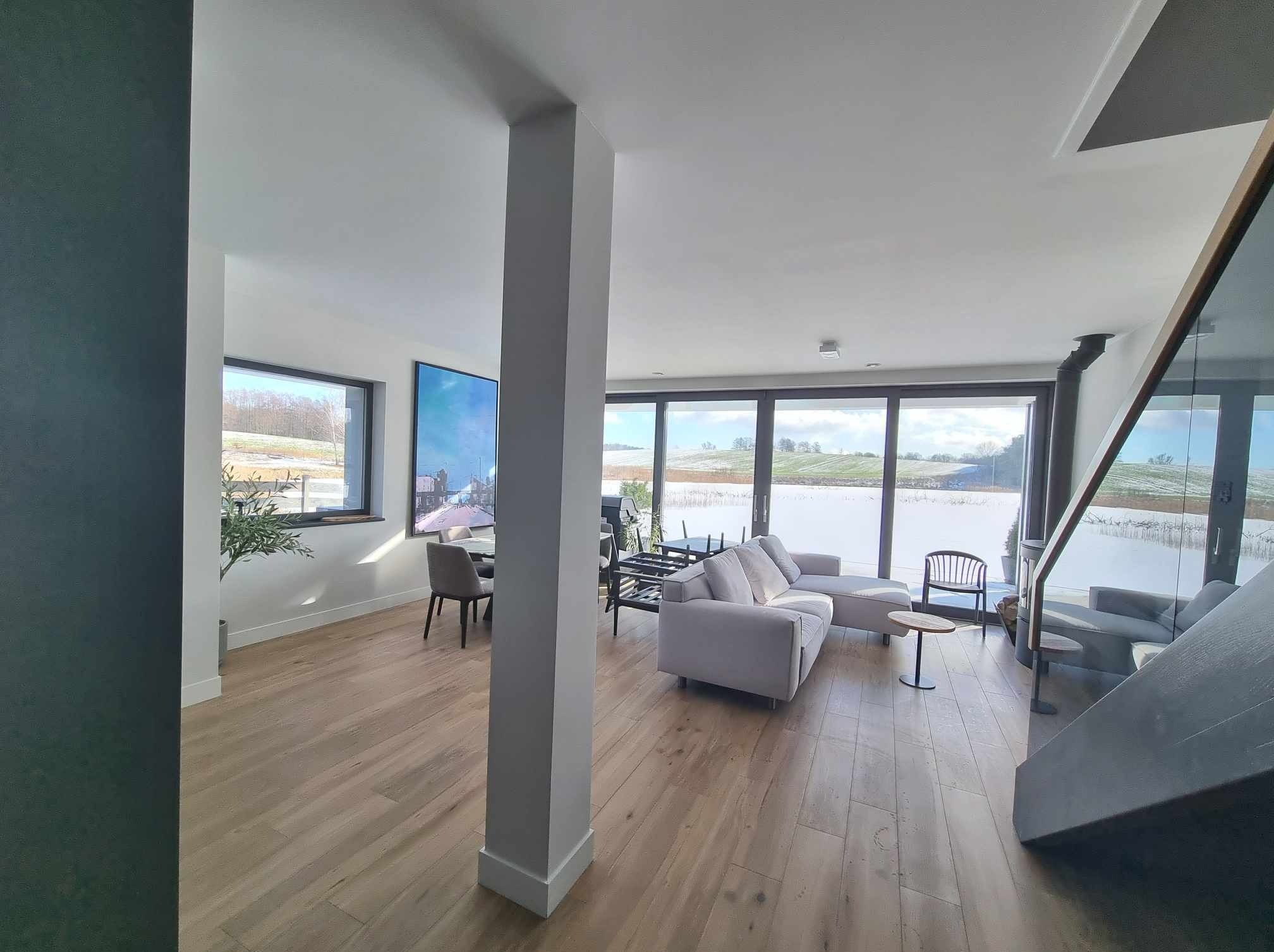 Jasne wnętrze domu z dużymi oknami, widok na zaśnieżony krajobraz, drewniana podłoga, szara sofa i kolumna.