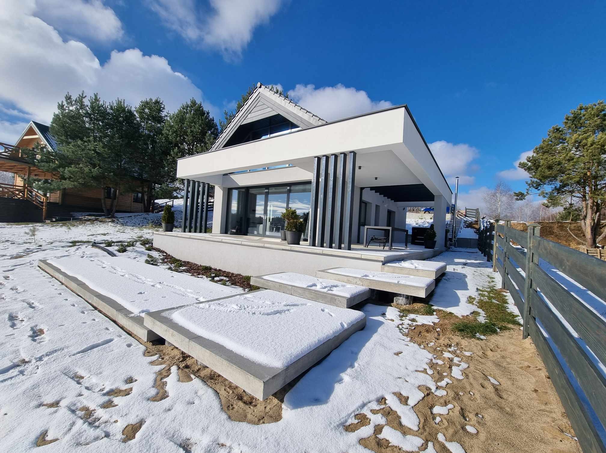 Nowoczesny, biały dom z płaskim dachem i czarnymi akcentami, otoczony zimowym krajobrazem z elementami betonowymi na pierwszym planie, częściowo pokrytymi śniegiem, widoczne drzewa iglaste w tle...