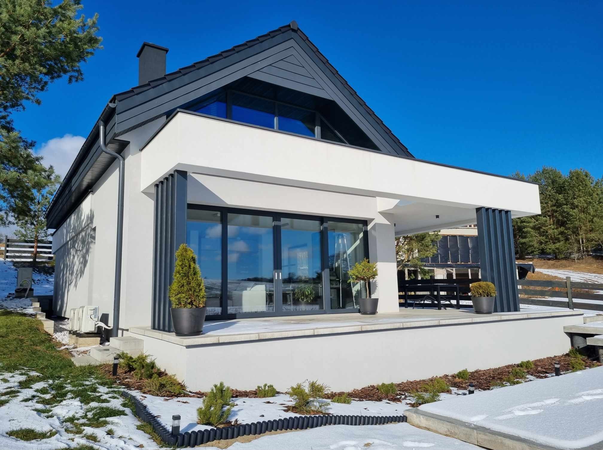 Nowoczesny dom z płaskim dachem i dużymi przeszkleniami, otoczony zimowym krajobrazem z fragmentami śniegu na trawniku i rabatach.