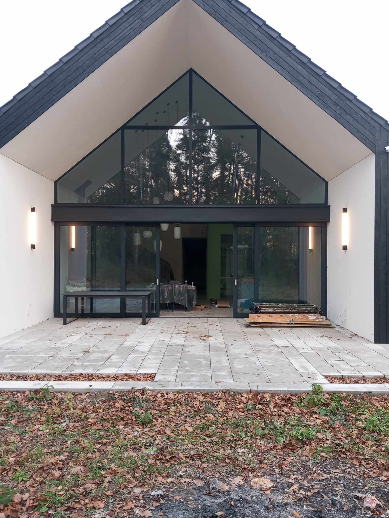 Nowoczesny dom z dużym trójkątnym oknem i przesuwnymi drzwiami szklanymi, oświetlony pionowymi lampami ściennymi, z wybrukowanym tarasem i widocznymi przygotowaniami do wykończenia wnętrza.