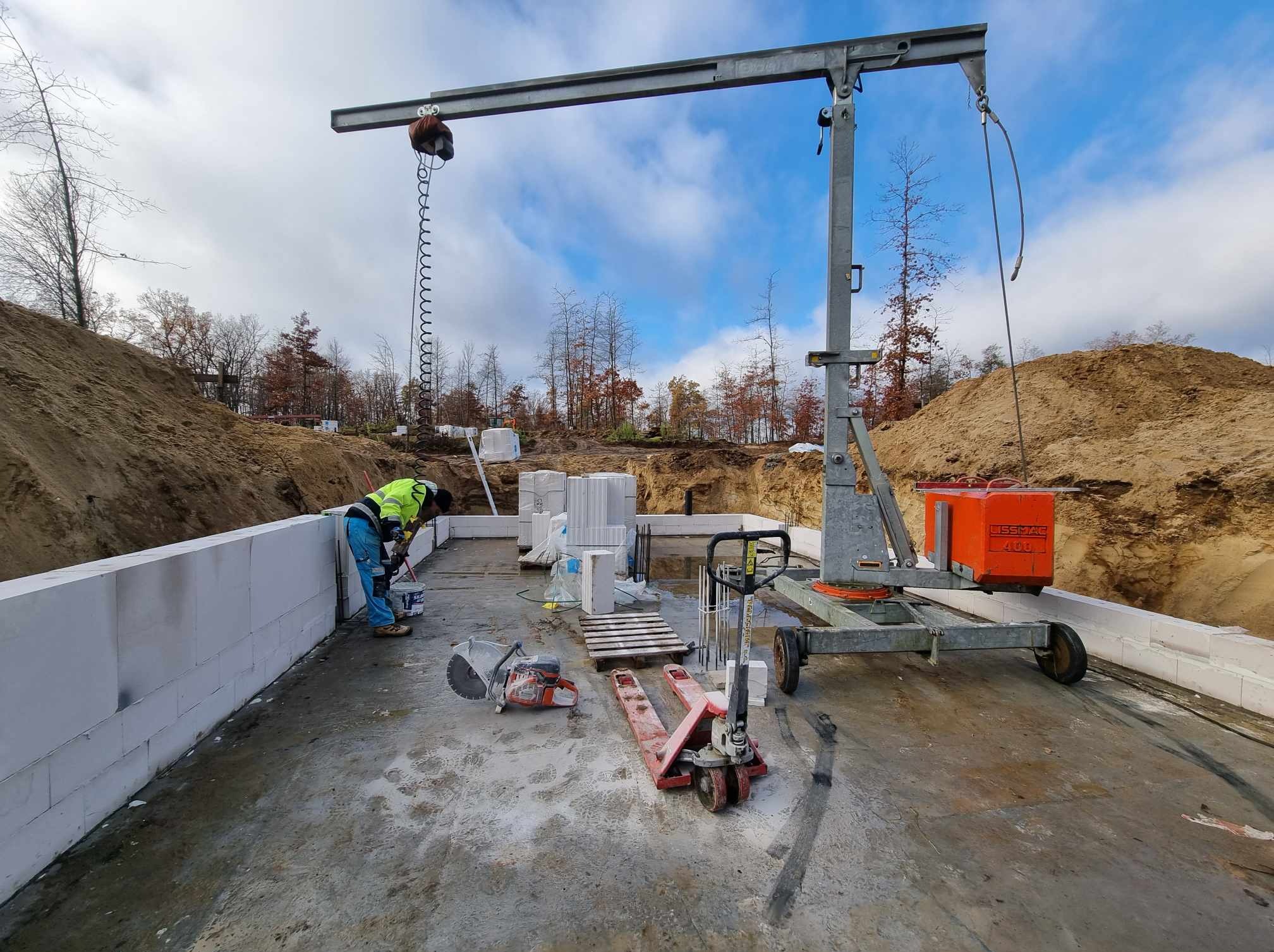 Budowa domu z bloczków: pracownik w odblaskowej kamizelce muruje ścianę fundamentową, widoczny dźwig, paleta z bloczkami, piła do cięcia bloczków oraz wózek paletowy.