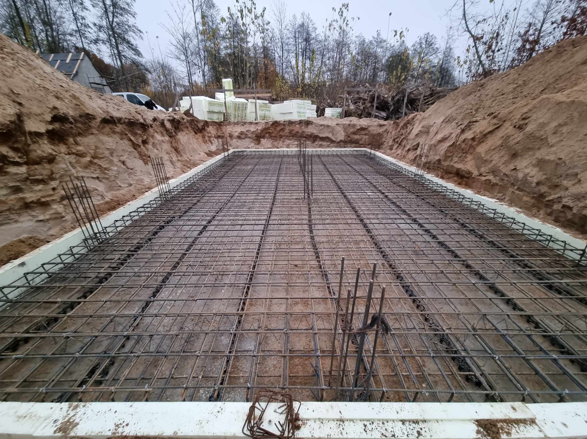 Przygotowany zbrojony szkielet fundamentu na gruncie, otoczony izolacją i wykopem ziemnym, z hałdą ziemi i stertą styropianu w tle.