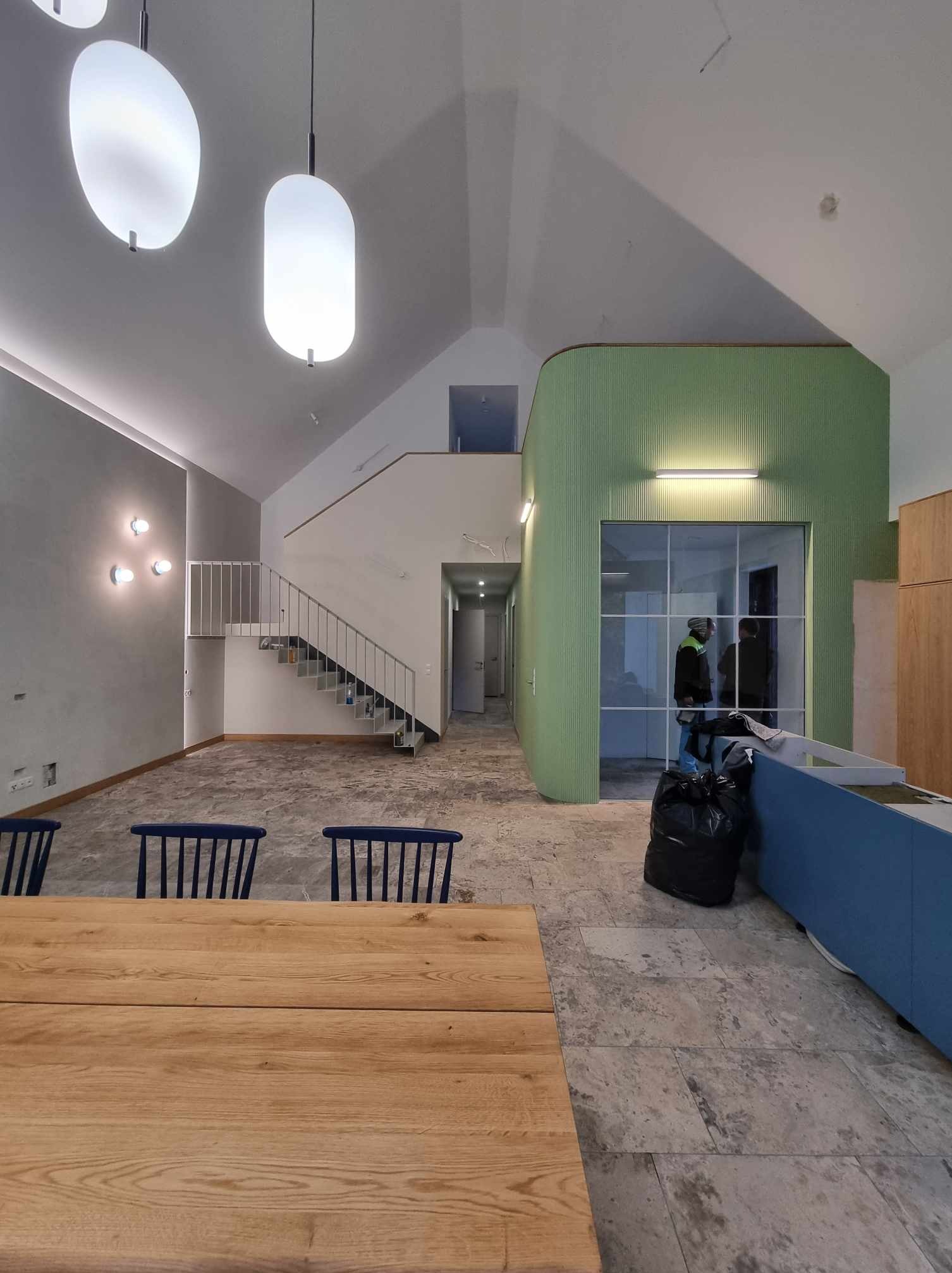 Wnętrze domu w stanie deweloperskim z drewnianym stołem na pierwszym planie, widoczne schody, surowe ściany i elementy wykończeniowe w odcieniach szarości i zieleni.