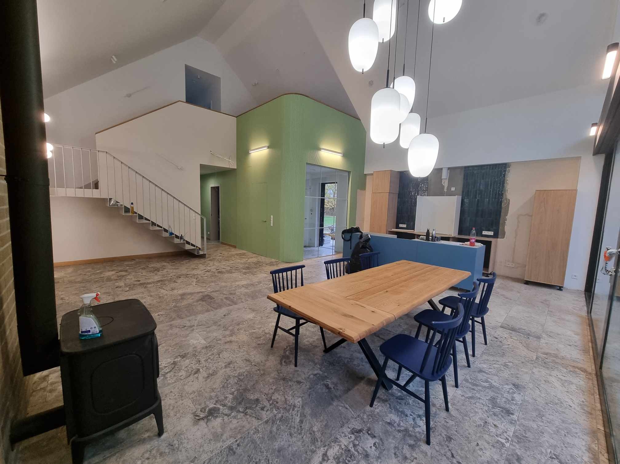 Wnętrze domu w trakcie wykańczania: surowa podłoga z szarego kamienia, drewniany stół z niebieskimi krzesłami, schody z metalową balustradą, piec wolnostojący i wiszące lampy w kształcie kul.