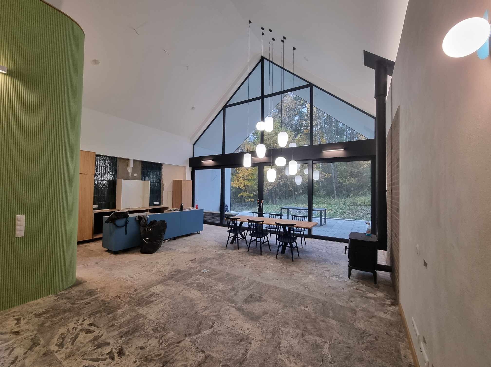 Wnętrze domu w trakcie wykańczania: widoczne duże okna z widokiem na zewnątrz, surowa podłoga z kamienia, niedokończone ściany i meble w tle, wiszące lampy o kulistym kształcie, czarny piec...