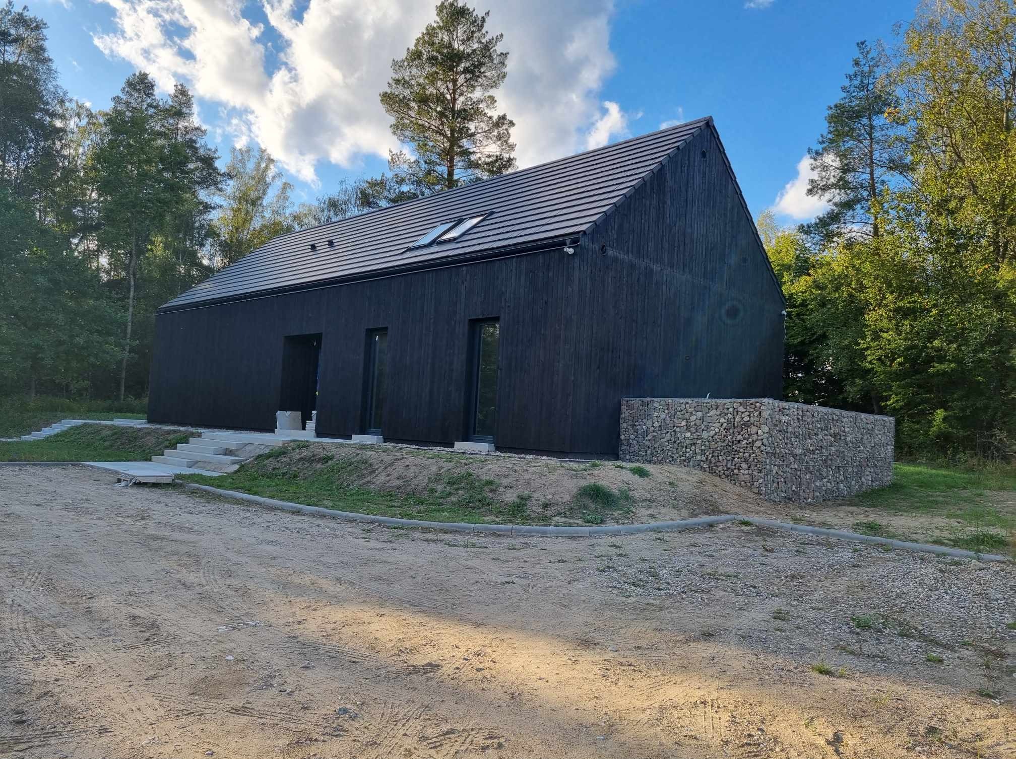 Nowoczesny dom murowany o czarnej elewacji z drewnianych desek, dwuspadowym dachem z dachówką i oknami dachowymi, otoczony zielenią i ziemią z widocznymi śladami opon.