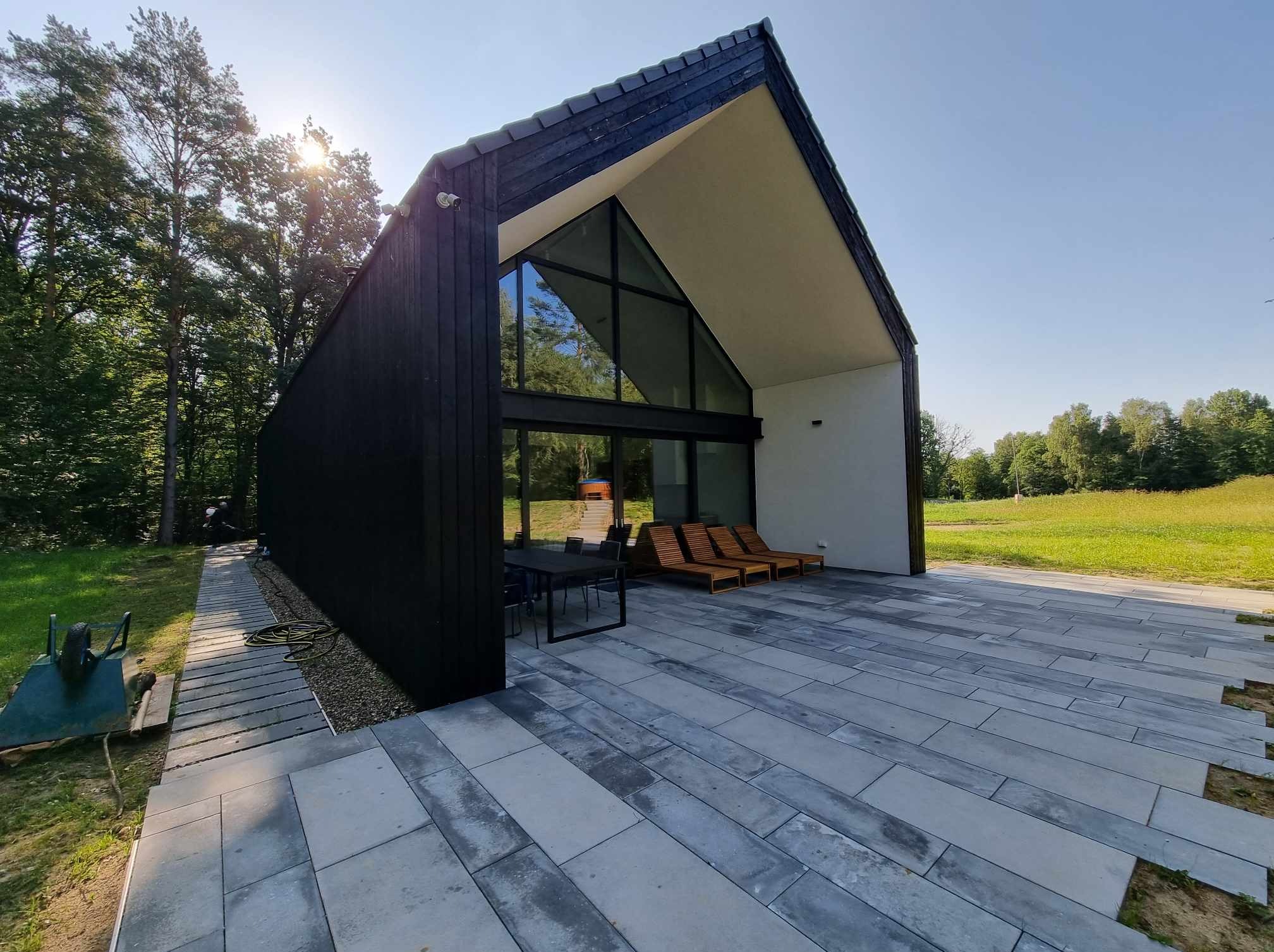 Nowoczesny dom z czarną drewnianą elewacją, dużymi przeszkleniami i tarasem wyłożonym szarymi płytami, otoczony zielenią drzew i trawnikiem.