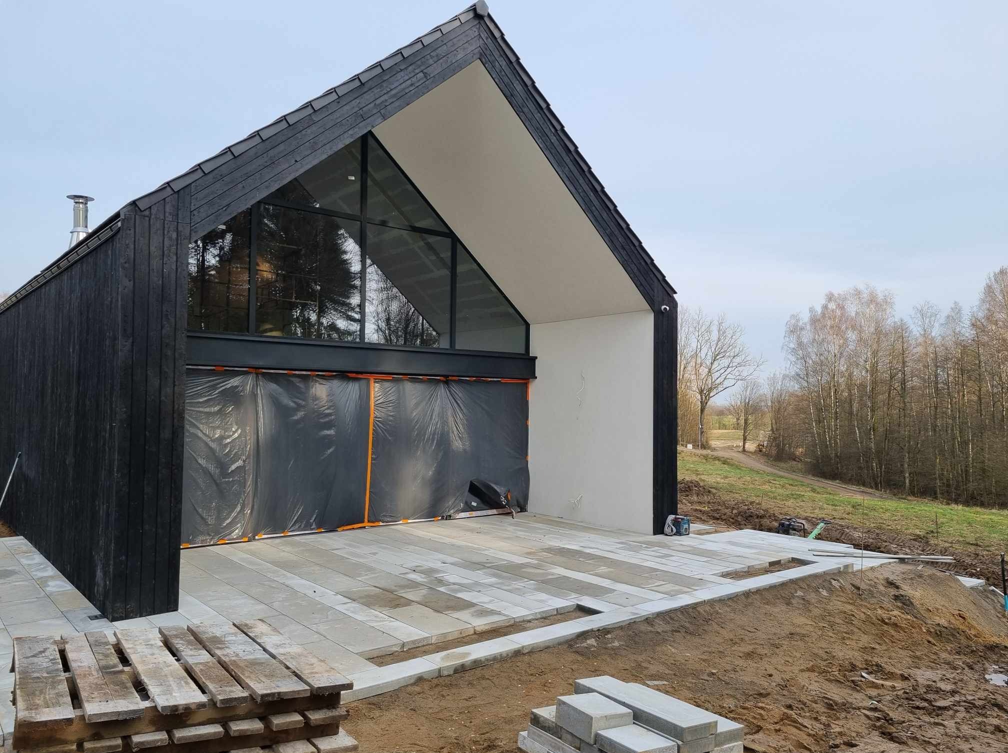 Nowoczesny dom z czarną drewnianą elewacją i dużym oknem trójkątnym, zabezpieczony folią budowlaną. Ułożona częściowo kostka brukowa na podjeździe, z paletą kostki obok. W tle widoczne drzewa.