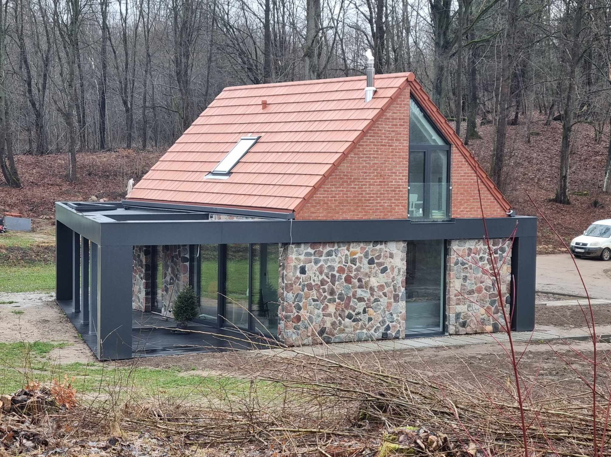 Nowoczesny dom z dachem z czerwonej dachówki, ceglaną górną kondygnacją i kamienną dolną częścią elewacji, z przeszklonymi ścianami i czarną konstrukcją tarasu, otoczony lasem i gałęziami.
