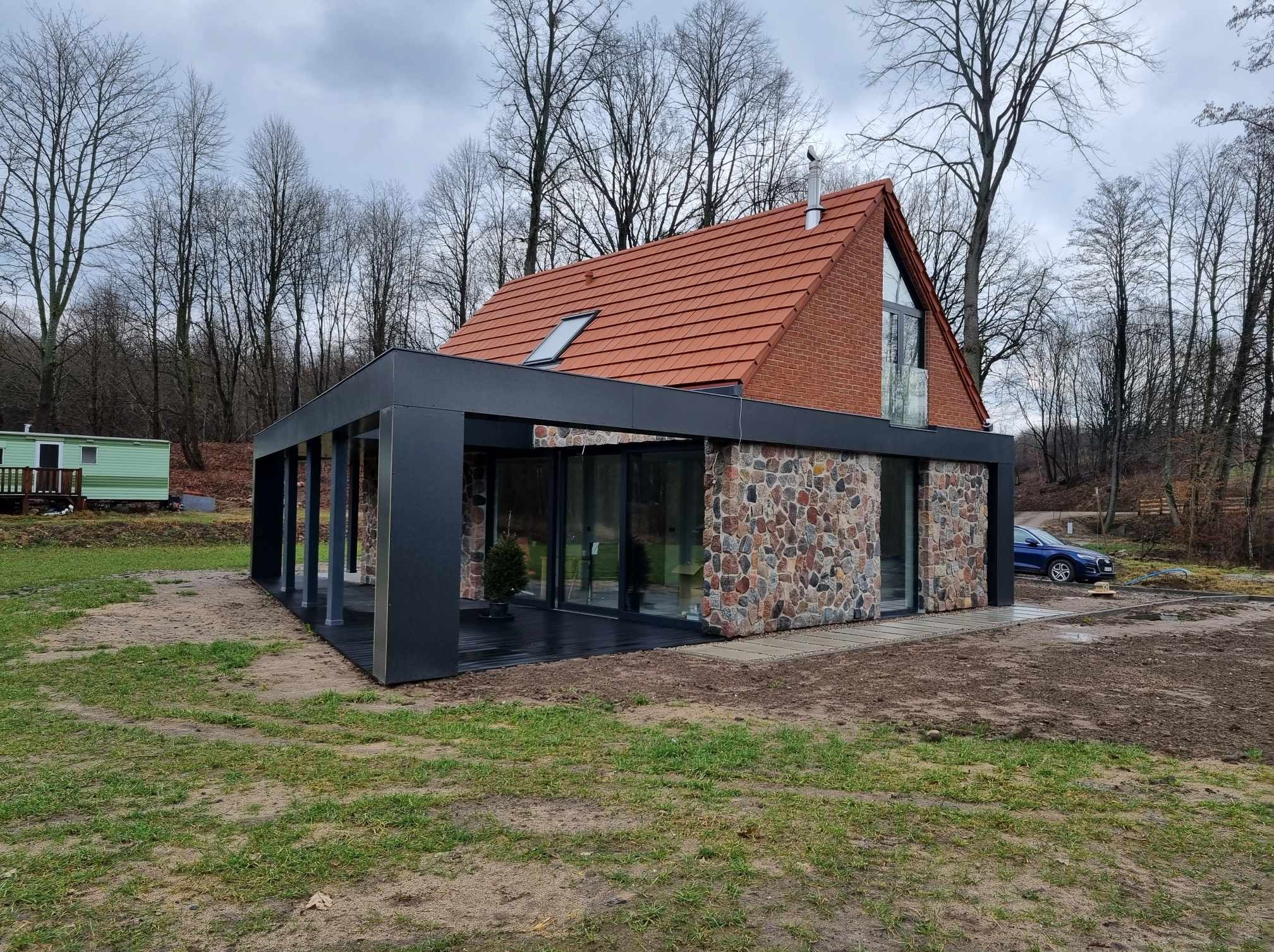 Nowoczesny dom jednorodzinny z czerwoną dachówką, elewacją z kamienia polnego i czarną, przeszkloną dobudówką, otoczony drzewami bez liści, widoczny samochód osobowy.