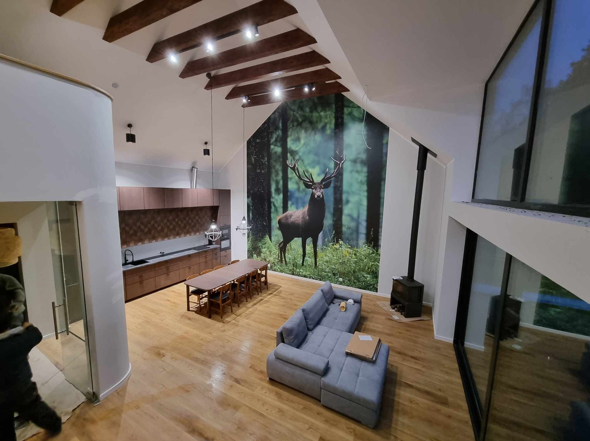 Przestronny salon z drewnianą podłogą, szarą sofą, stołem jadalnym i fototapetą z motywem jelenia w lesie, widok z góry.