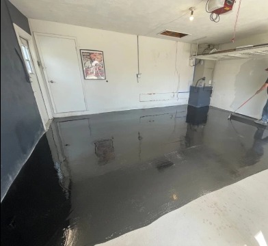 Aplikacja czarnej posadzki żywicznej w garażu. Widoczny pracownik rozprowadzający żywicę wałkiem, ściany garażu i oświetlenie. Błyszcząca, mokra powierzchnia.