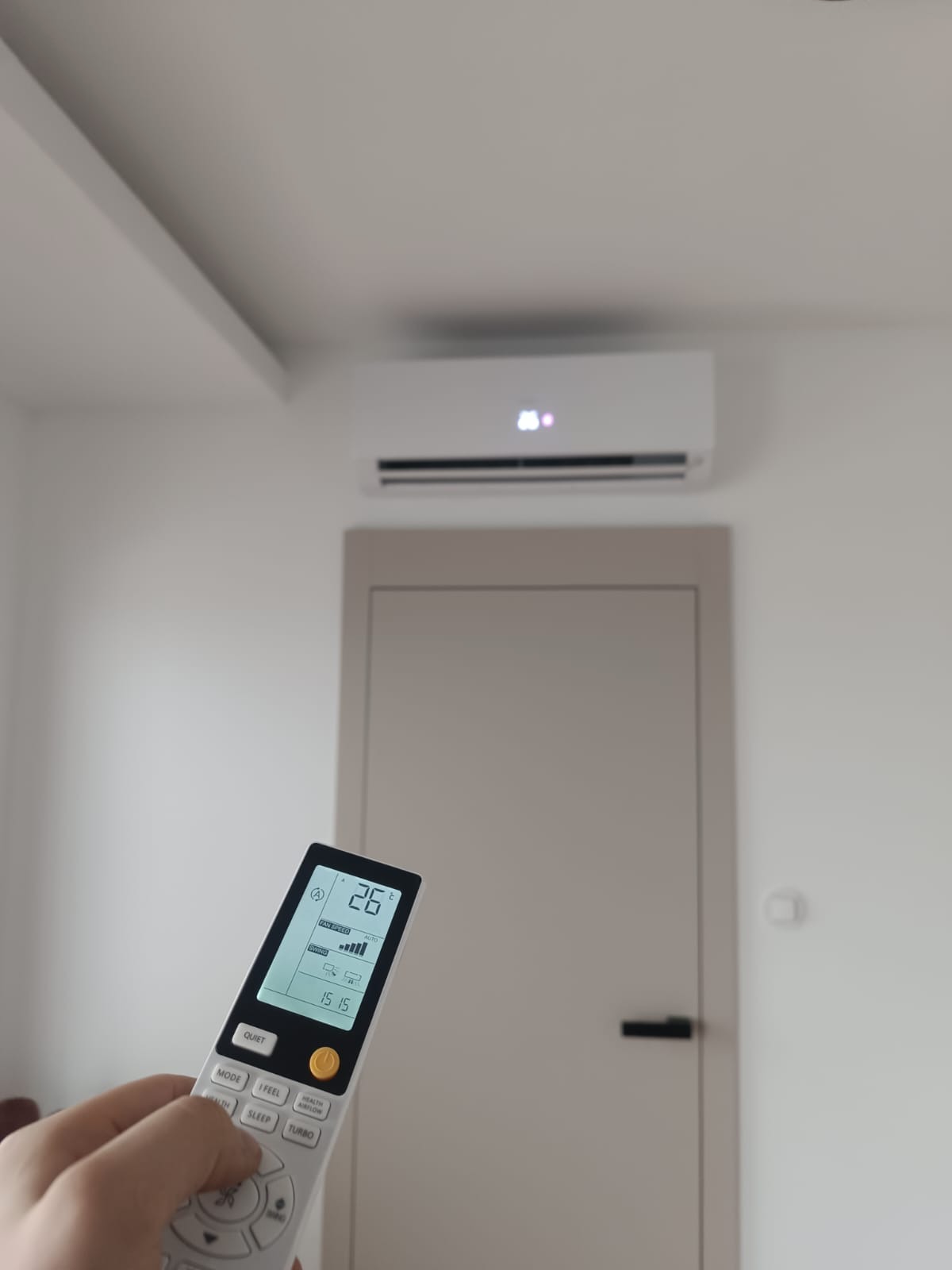 Biały klimatyzator ścienny włączony, ustawiony na 26 stopni Celsjusza, sterowany pilotem trzymanym w dłoni, na ścianie pomalowanej na biało, widoczny fragment sufitu i drzwi w tle.