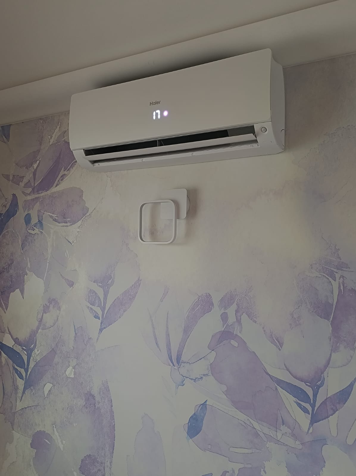 Biały klimatyzator Haier zamontowany na ścianie z delikatnym, kwiatowym wzorem w odcieniach fioletu i błękitu, wyświetlający temperaturę 17 stopni.