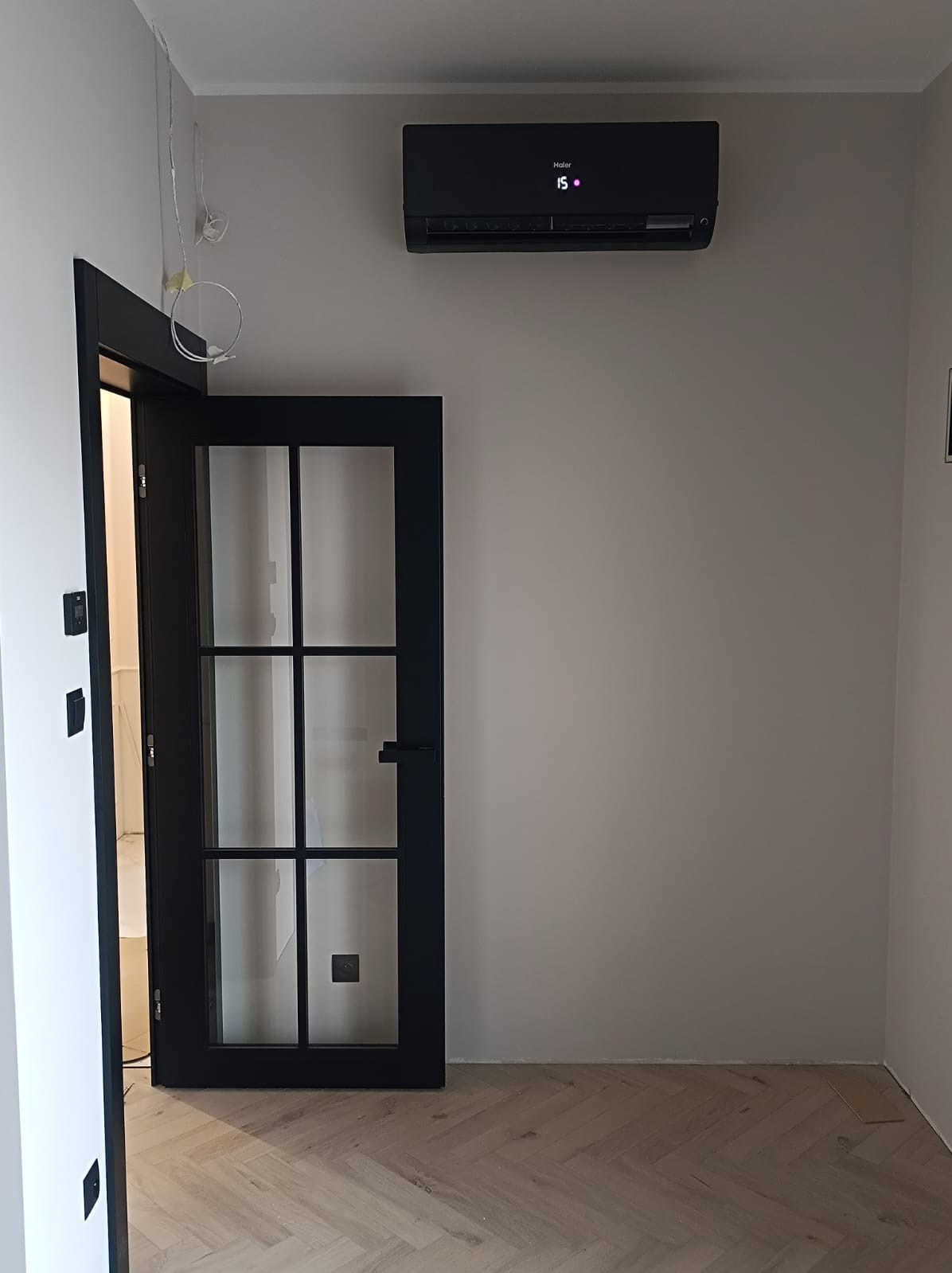 Czarny klimatyzator Haier zamontowany na ścianie nad otwartymi czarnymi drzwiami w nowocześnie wykończonym wnętrzu z parkietem w jodełkę.