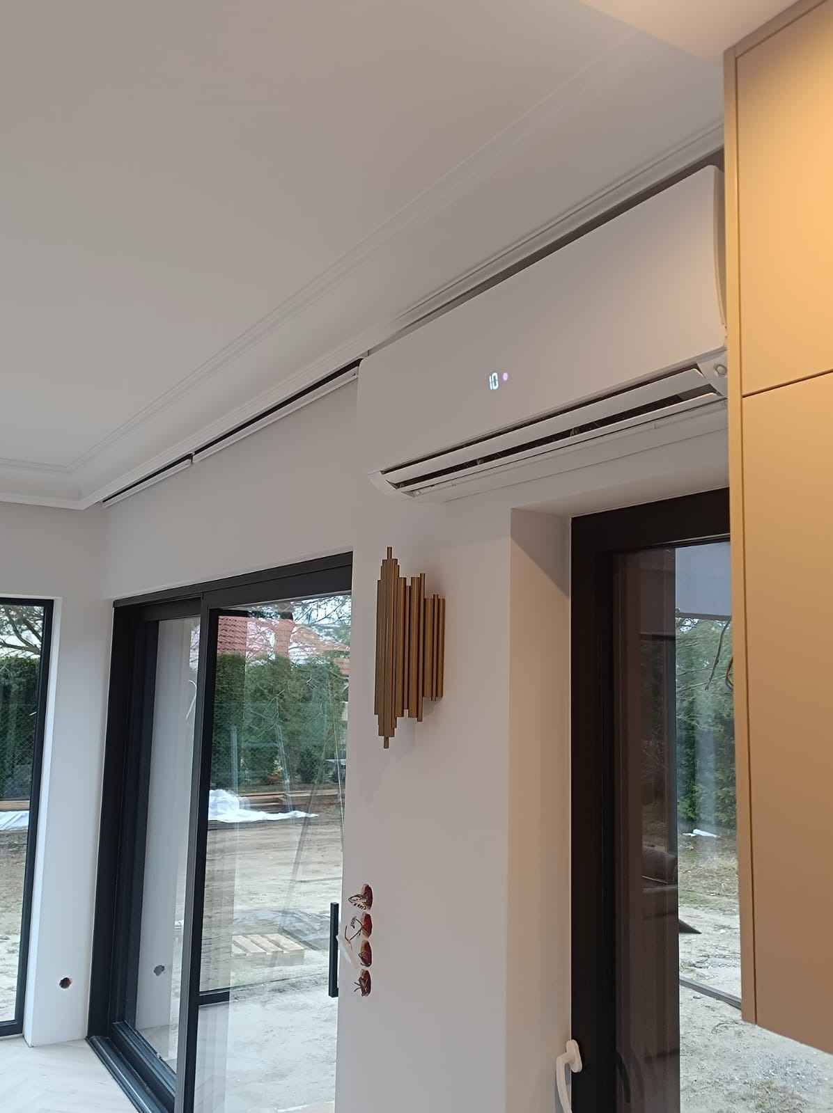 Biały klimatyzator ścienny z wyświetlaczem temperatury w nowoczesnym wnętrzu, widoczne ozdobne listwy sufitowe i złota dekoracja ścienna, duże okno przesuwne z czarną ramą.