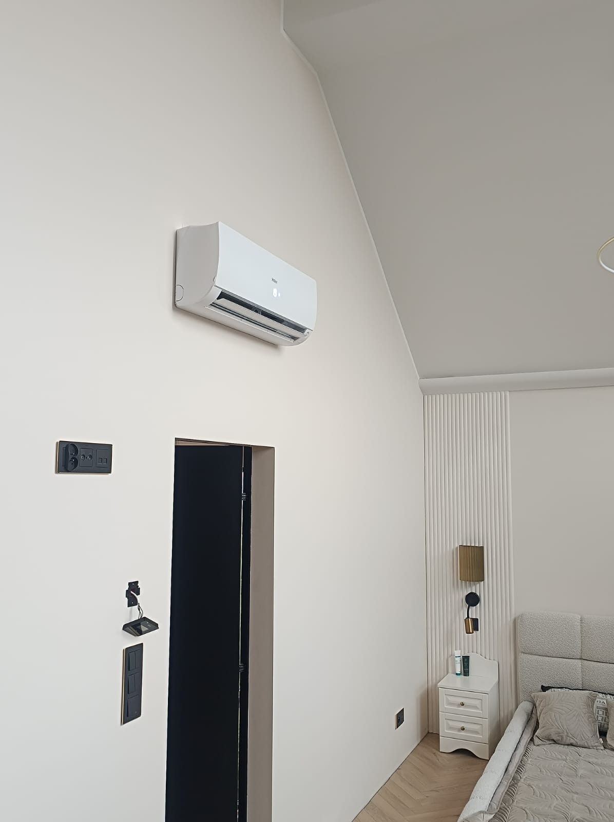 Biały klimatyzator Haier zamontowany na ścianie w jasnym pokoju z czarnymi włącznikami i gniazdkami, widoczny fragment sypialni z łóżkiem i stolikiem nocnym.