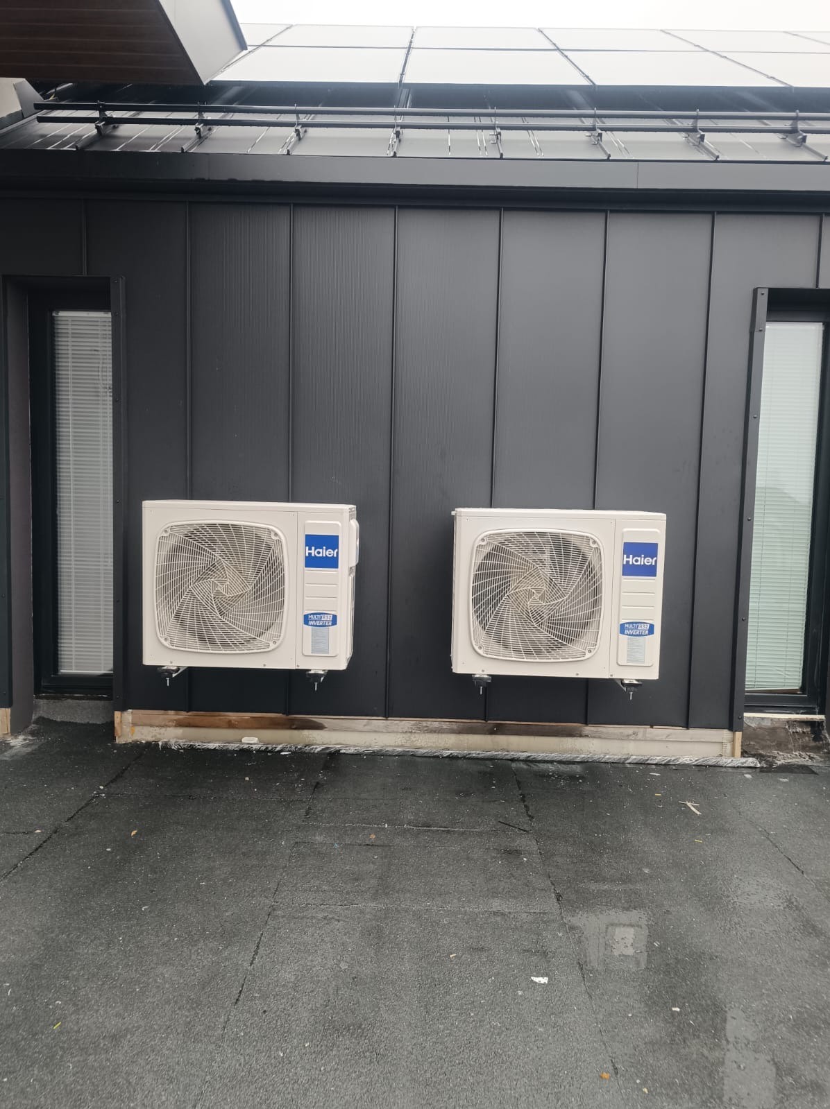 Dwa białe klimatyzatory marki Haier zamontowane na ciemnej, metalowej elewacji budynku, widoczne okna z żaluzjami i mokry chodnik.