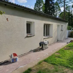 Elewacja budynku po nałożeniu tynku strukturalnego, widoczne okna i drzwi. Przy budynku stoi wiadro z zaprawą. Tło stanowią drzewa i błękitne niebo.