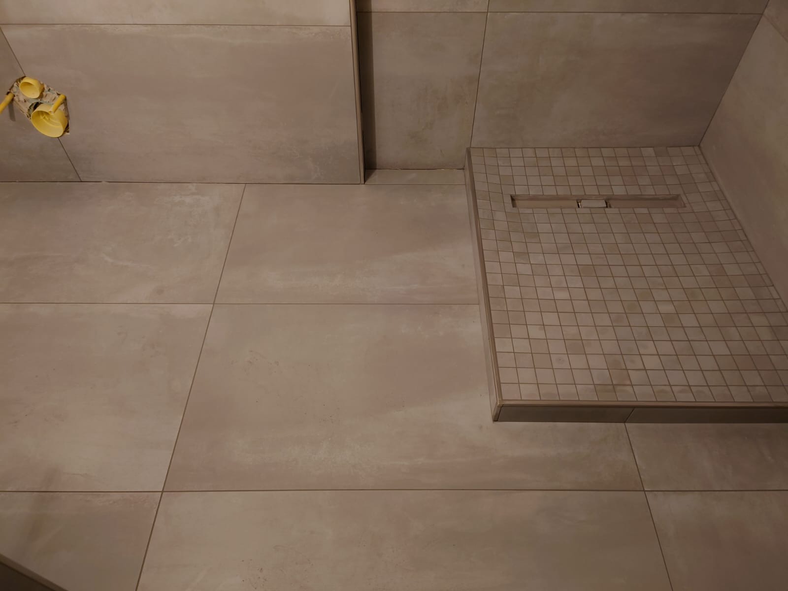 Beżowe, matowe płytki w łazience z odpływem liniowym w kabinie prysznicowej. Widoczne żółte elementy instalacji wodnej. Minimalistyczny design.