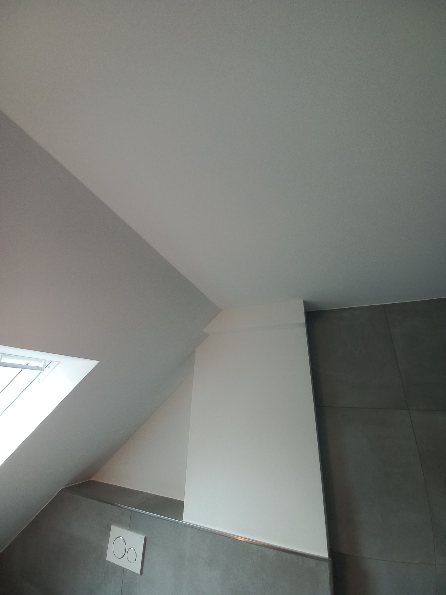 Wykończona łazienka na poddaszu w Nakle nad Notecią. Szare płytki, białe ściany i okno dachowe. Widoczny przycisk spłuczki. Minimalistyczny design.