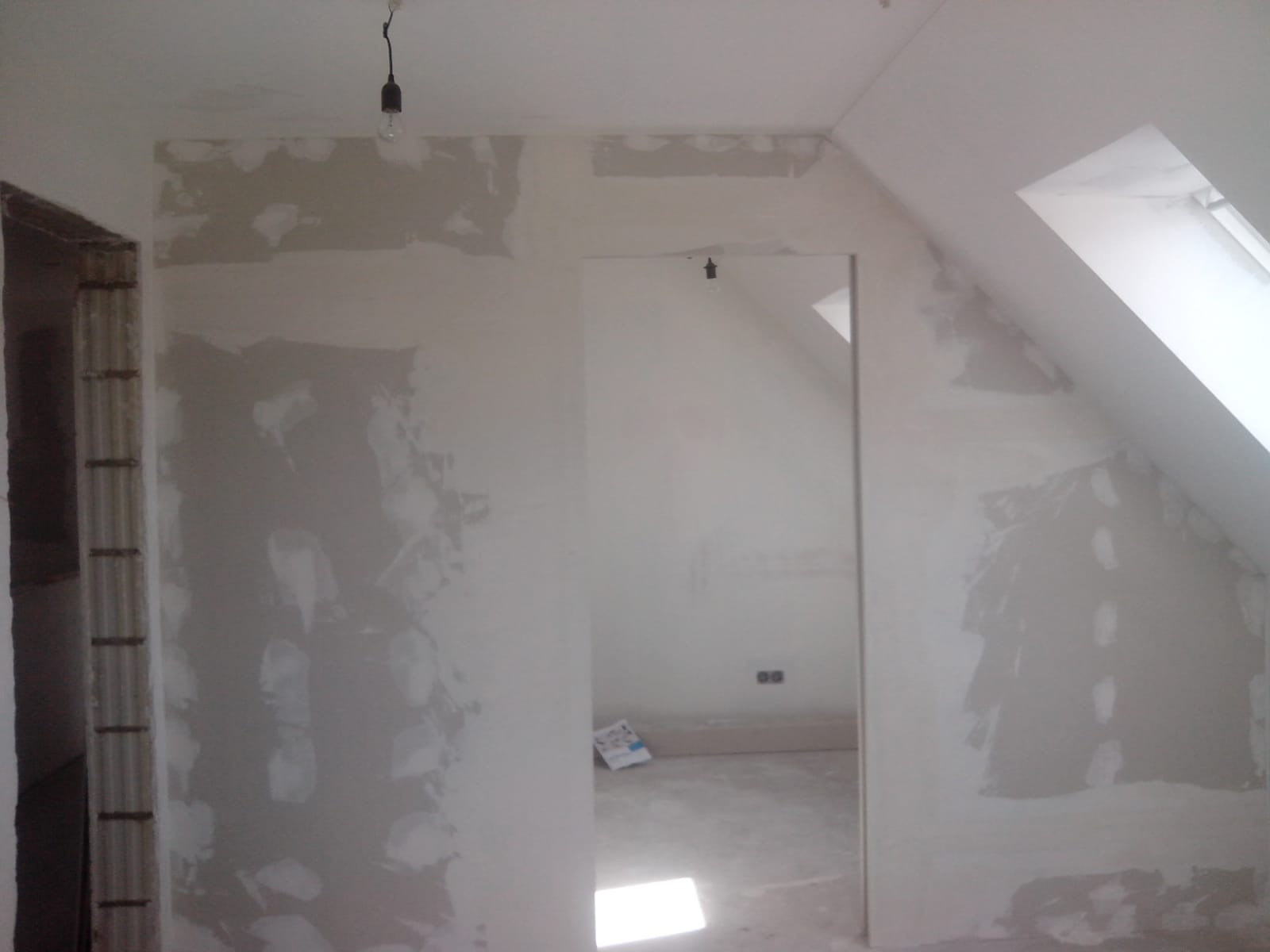 Poddasze w trakcie prac wykończeniowych. Ściany częściowo pokryte gładzią, widoczne ślady po szpachlowaniu. Surowa żarówka zwisa z sufitu. Okno dachowe w skosie.