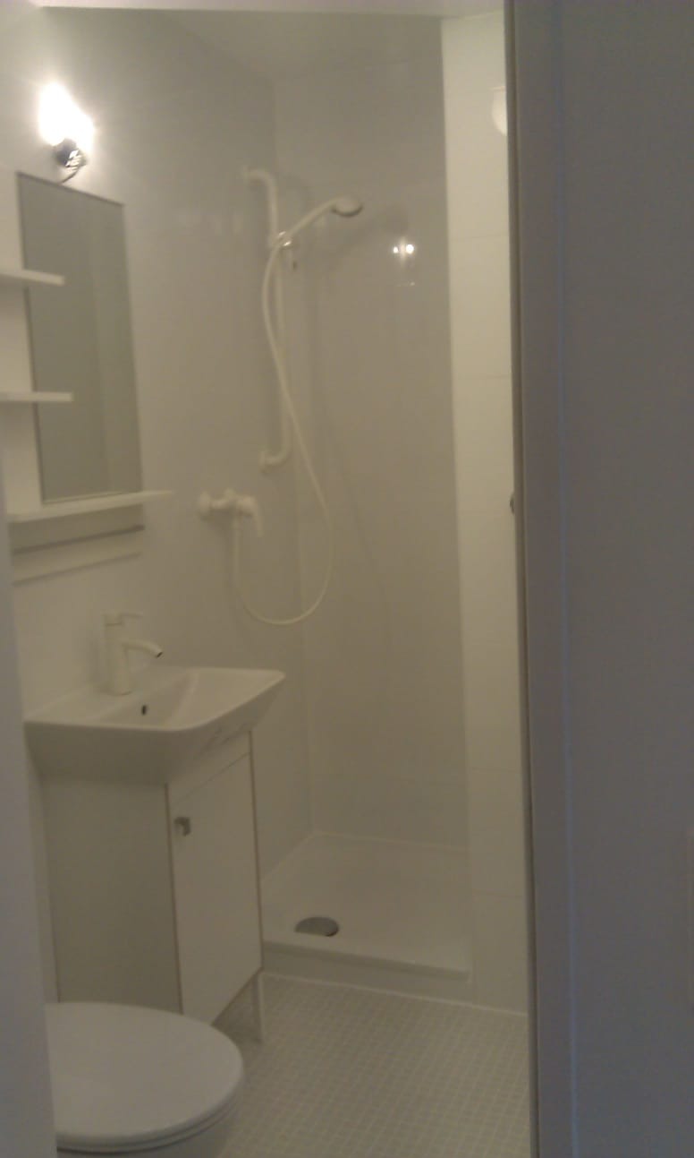 Jasna, minimalistyczna łazienka z kabiną prysznicową typu walk-in, umywalką z szafką i toaletą. Białe płytki podłogowe i ścienne. Nowoczesne oświetlenie.