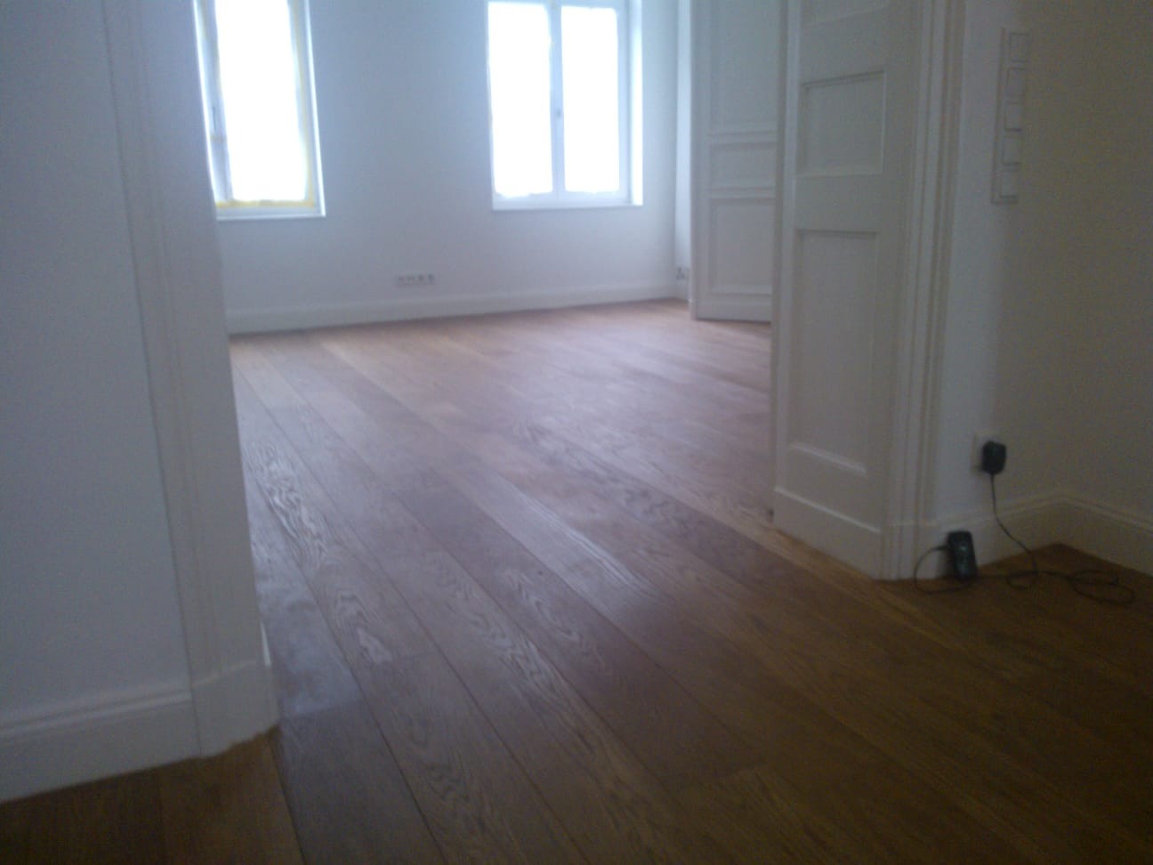 Jasne, przestronne wnętrze po remoncie: drewniana podłoga z paneli, białe ściany i stolarka okienna. Widoczne gniazdka elektryczne przy podłodze.