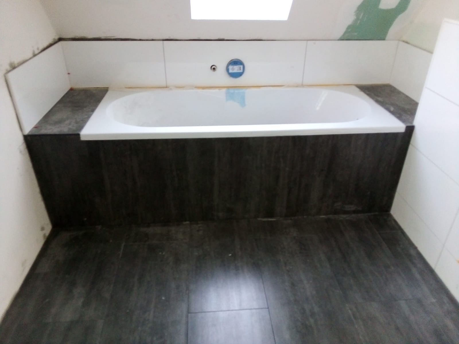 Biała wanna obudowana ciemnymi płytkami imitującymi drewno, z białymi płytkami na ścianie w łazience z oknem dachowym. Widoczne fugi i fragmenty niezakończonych prac.