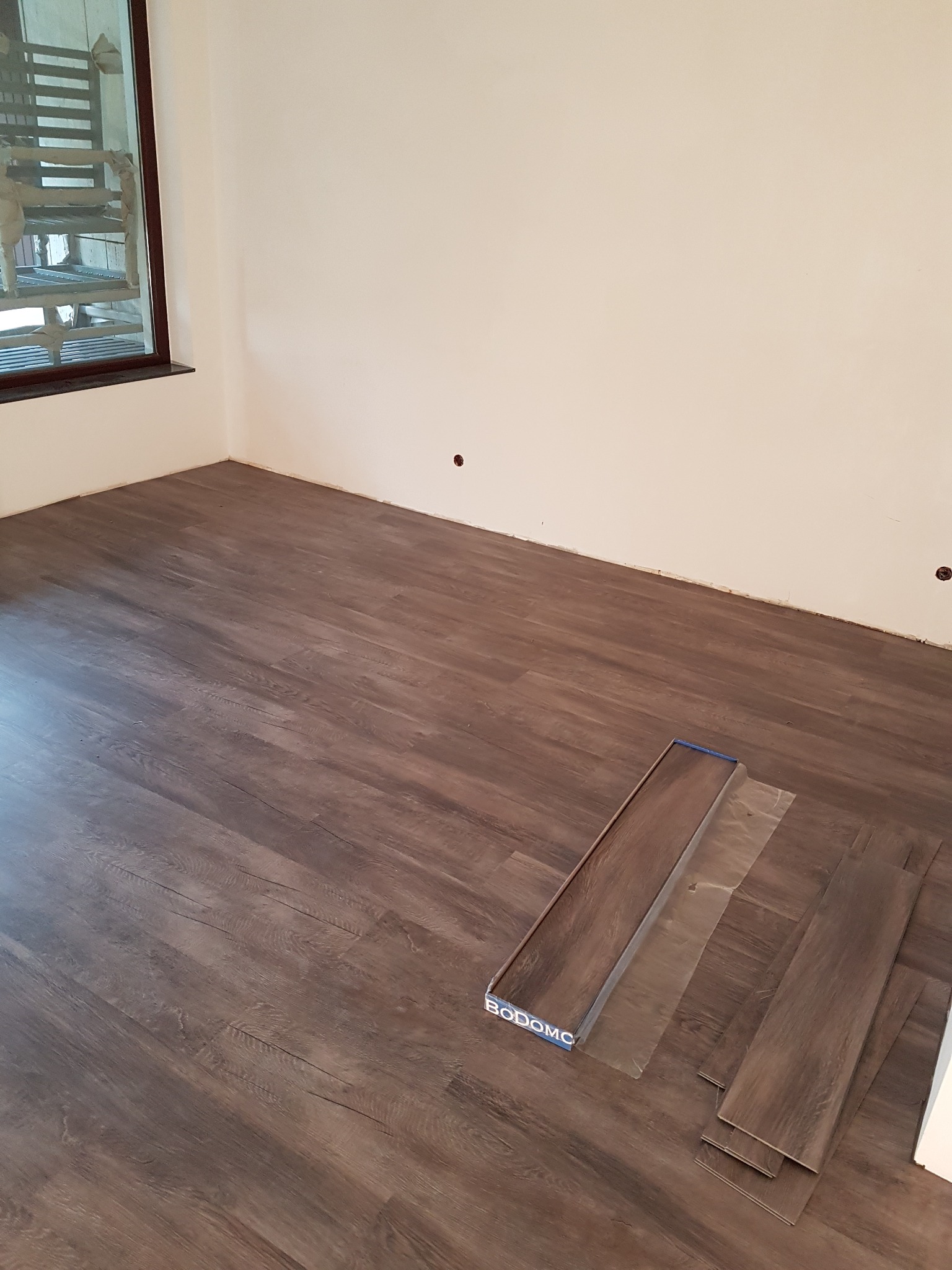 Układanie paneli podłogowych w pokoju, widoczne panele marki BODOMC, brązowe, drewnopodobne, częściowo ułożone, ściany białe, okno w lewym górnym rogu.