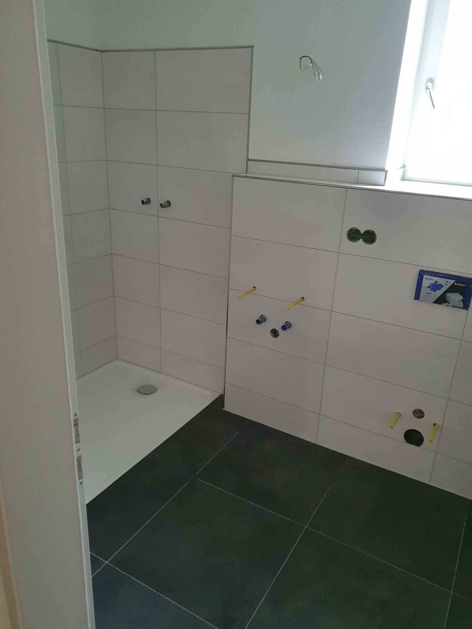 Nowoczesna łazienka w trakcie wykańczania: białe płytki ścienne, ciemna podłoga, przygotowane instalacje wod-kan, widoczny brodzik. Minimalistyczny design.