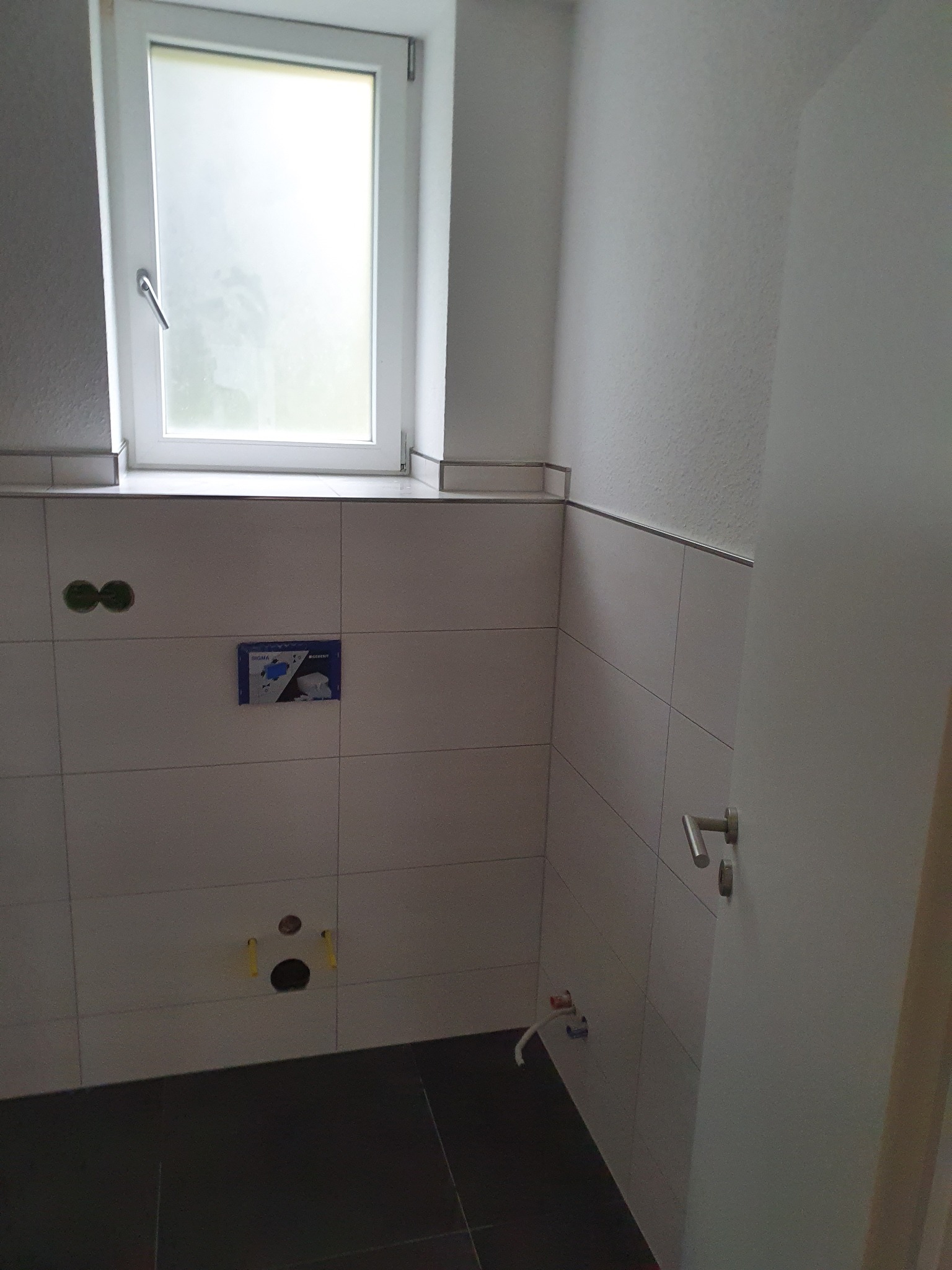 Wykończona łazienka z białymi płytkami ściennymi, ciemną podłogą i oknem. Widoczne przygotowane punkty instalacyjne i fragment drzwi z klamką.