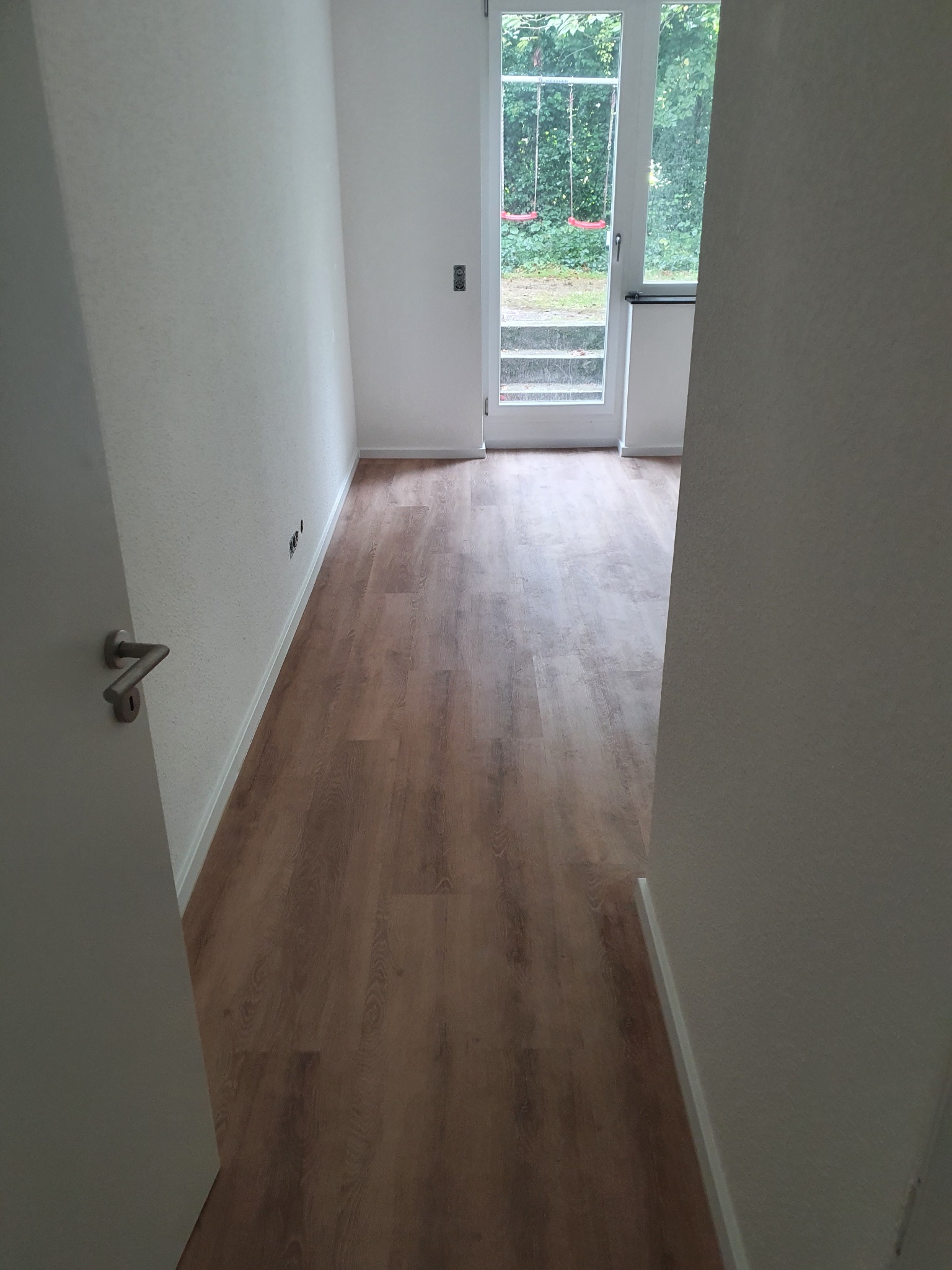 Jasne panele podłogowe w nowym wnętrzu, widok zza otwartych drzwi. Świeżo pomalowane ściany i widok na ogród z huśtawką przez przeszklone drzwi balkonowe.