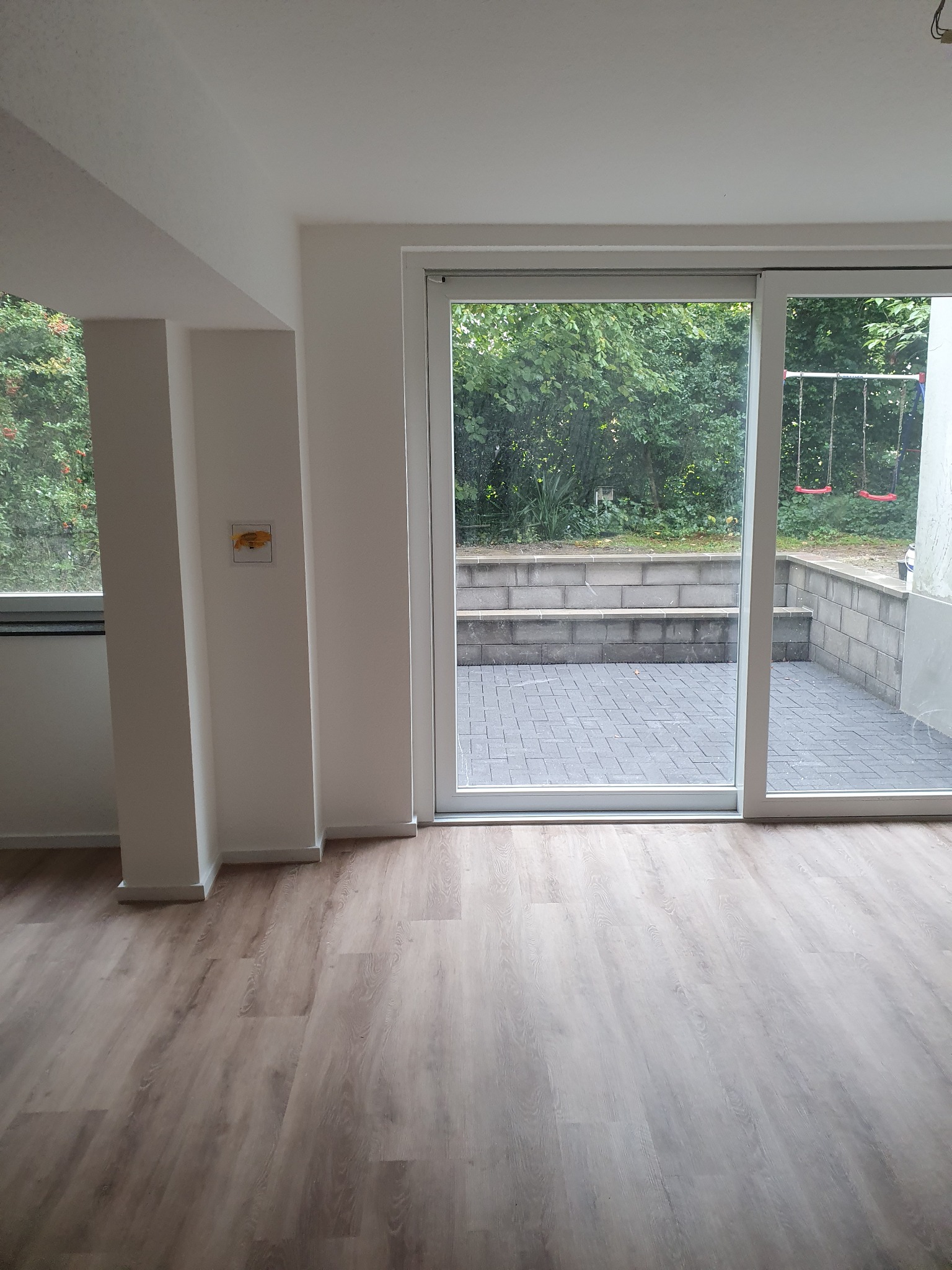 Jasne panele podłogowe w nowym wnętrzu z widokiem na taras i plac zabaw przez duże okno przesuwne. Minimalistyczny design, świeżo po remoncie.