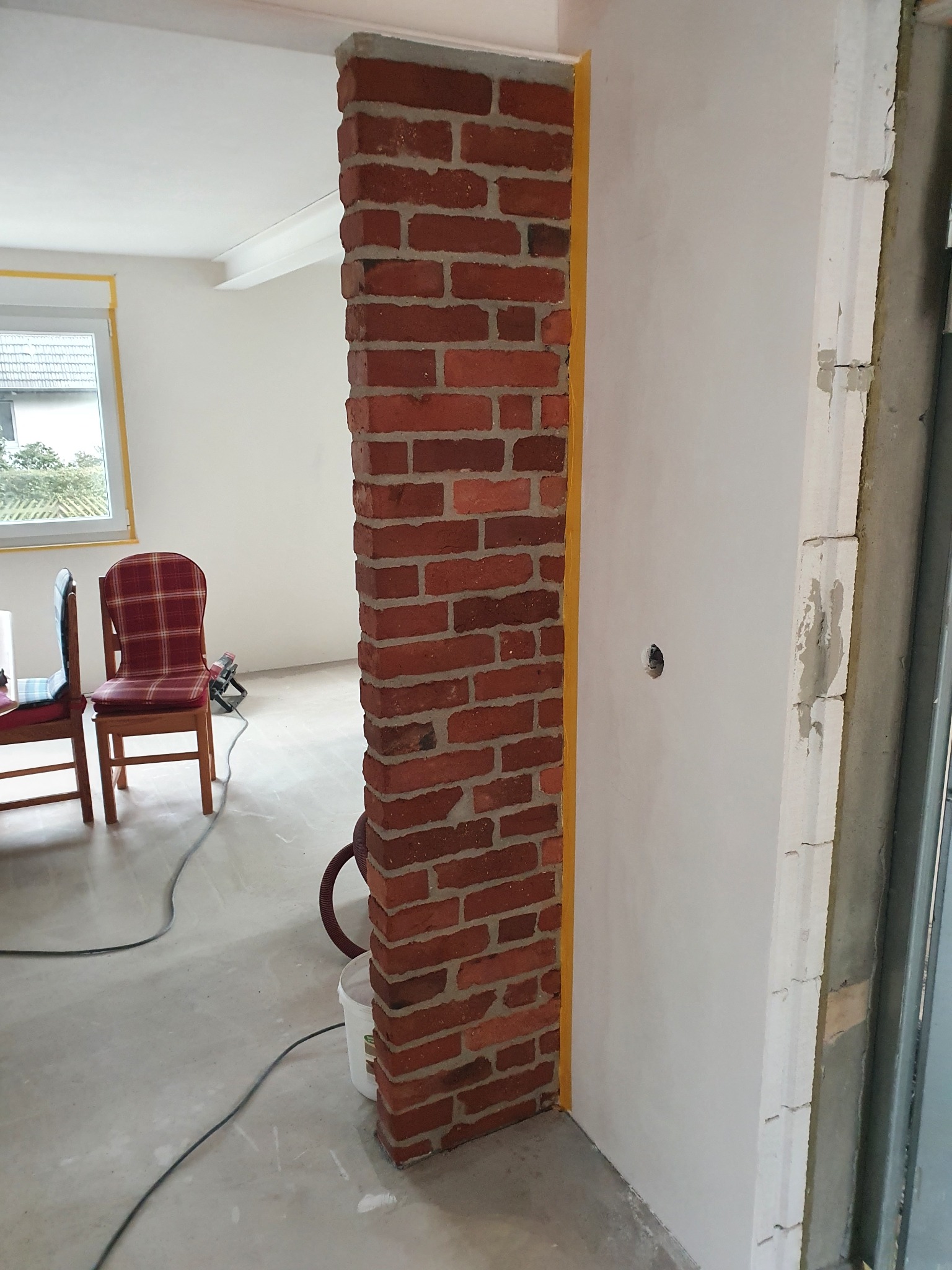 Ceglana kolumna z żółtą taśmą maskującą, świeżo otynkowana ściana w tle, widoczne krzesła i sprzęt budowlany. Etap prac wykończeniowych w pomieszczeniu.