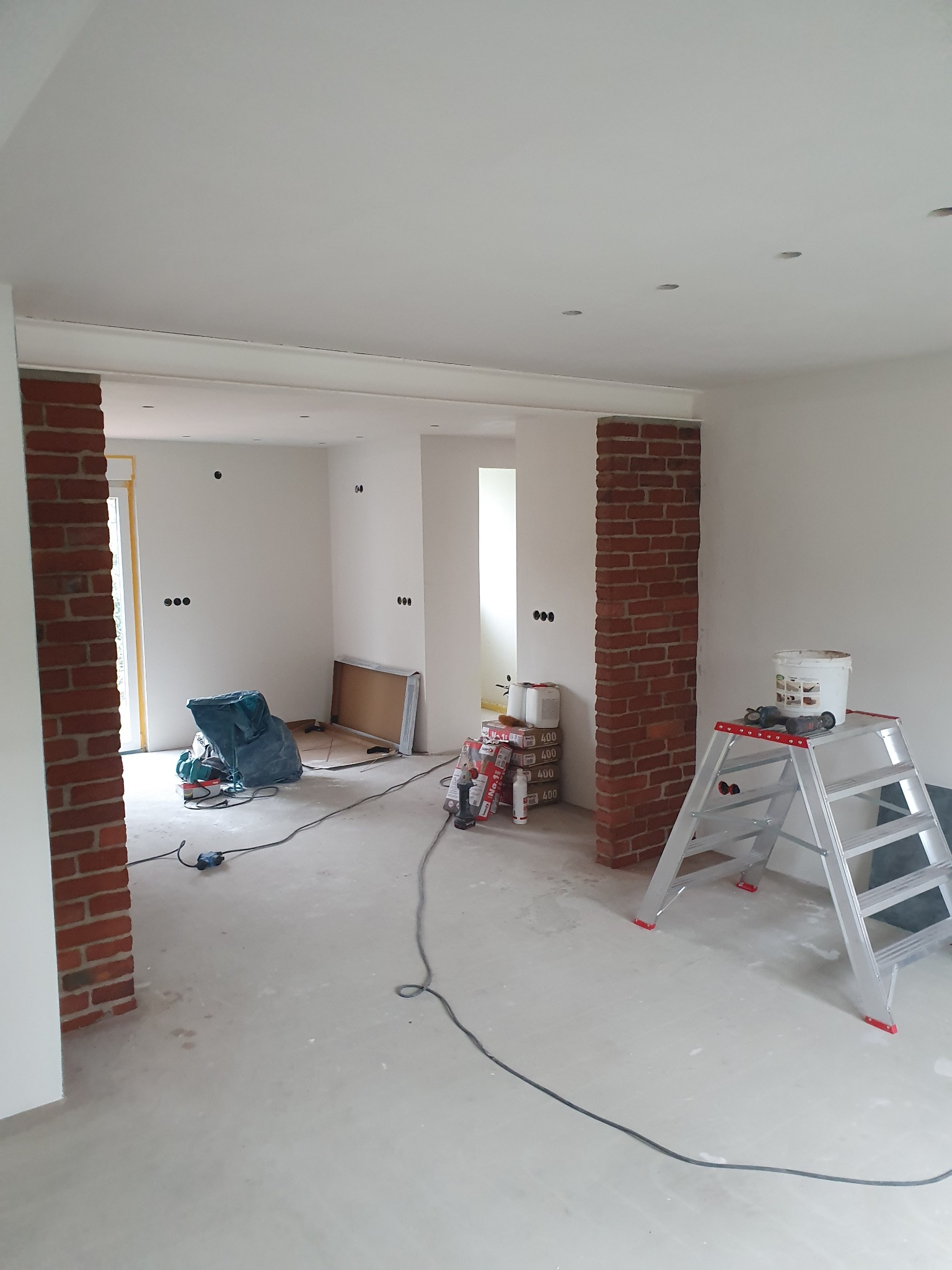 Wnętrze w trakcie prac wykończeniowych: surowe ściany, ceglane filary, drabina malarska, narzędzia i materiały budowlane. Widoczne puszki elektryczne.