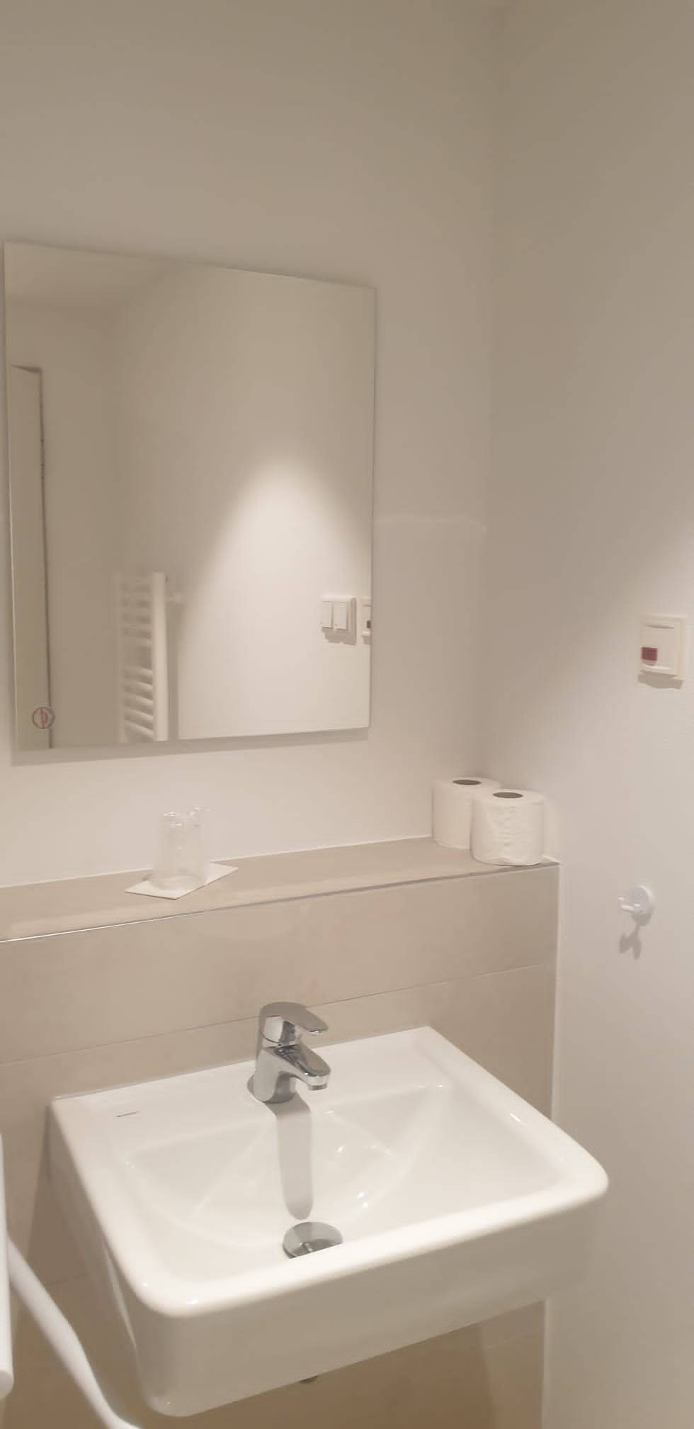 Nowoczesna, minimalistyczna łazienka z białą umywalką, chromowaną baterią i lustrem z podświetleniem LED. Widoczne akcesoria łazienkowe i grzejnik drabinkowy w odbiciu.