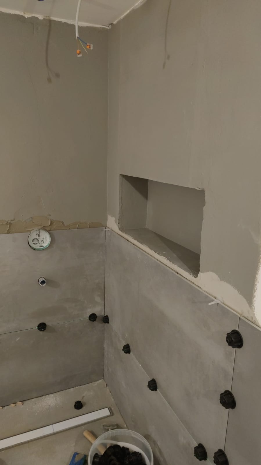 Szare płytki ścienne w trakcie montażu w łazience, z widoczną wnęką i elementami poziomowania. Ujęcie z dołu, z widocznym odpływem liniowym i wiadrem z akcesoriami.