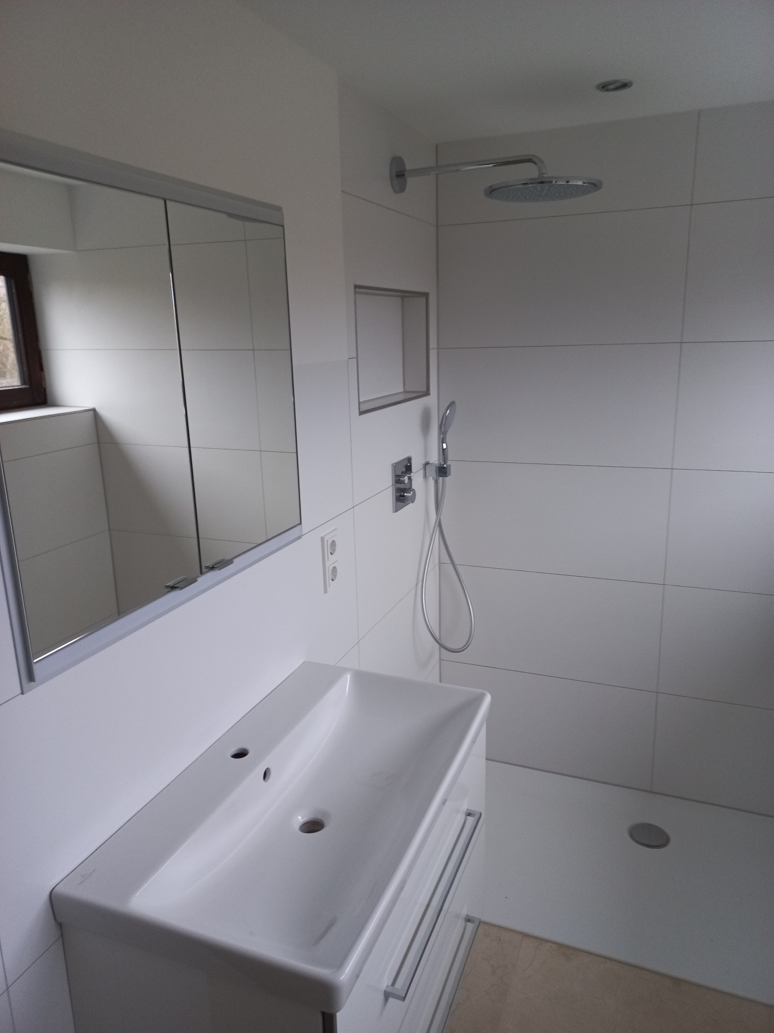 Nowoczesna, minimalistyczna łazienka z białymi płytkami, kabiną prysznicową typu walk-in i umywalką z szafką. Lustro z szafką w tle. Widoczne detale armatury.