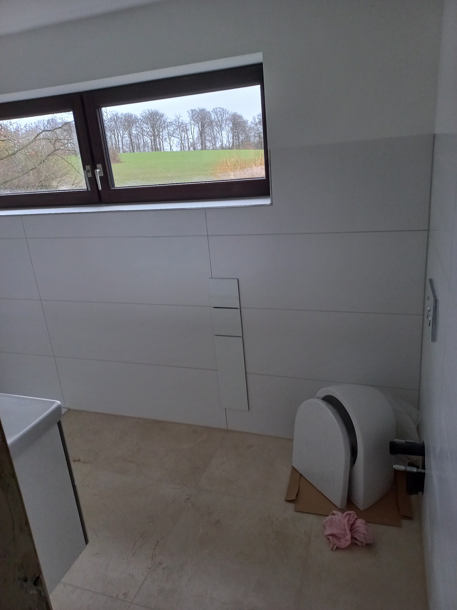 Nowoczesna łazienka w trakcie wykańczania: białe płytki imitujące panele, okno z widokiem na zieleń, częściowo zamontowana toaleta podtynkowa.