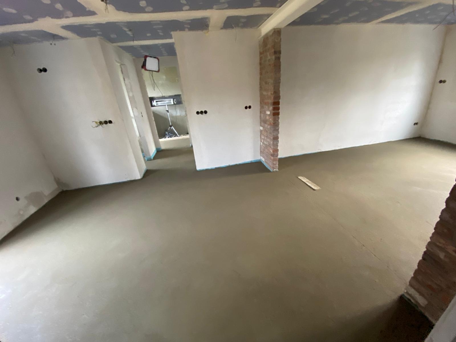 Wnętrze w trakcie prac wykończeniowych: wylana posadzka, ściany przygotowane pod gładzie, widoczne instalacje elektryczne. Fragment ściany z cegły.