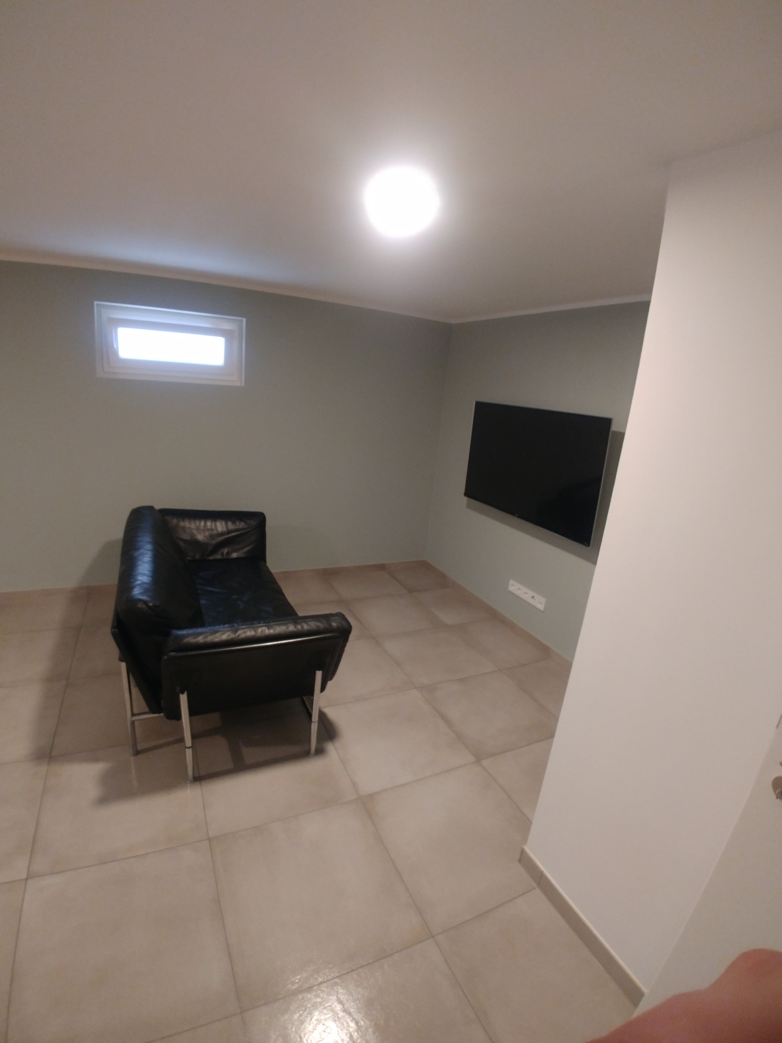 Salon po remoncie: czarna skórzana sofa na tle świeżo pomalowanych ścian w odcieniach szarości i beżu, z zamontowanym telewizorem. Jasne płytki podłogowe.