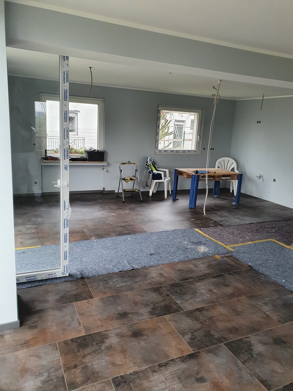 Wnętrze w trakcie remontu: brązowe płytki podłogowe, zabezpieczone fragmenty podłogi, szare ściany, okna z widokiem na zieleń, drabina i krzesła w tle. Widoczne kable ze stropu.