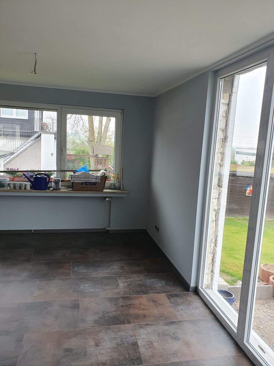 Pomieszczenie po malowaniu ścian na szaro i ułożeniu brązowych paneli podłogowych. Widoczne okno z widokiem na zewnątrz i fragment ściany z grzejnikiem.