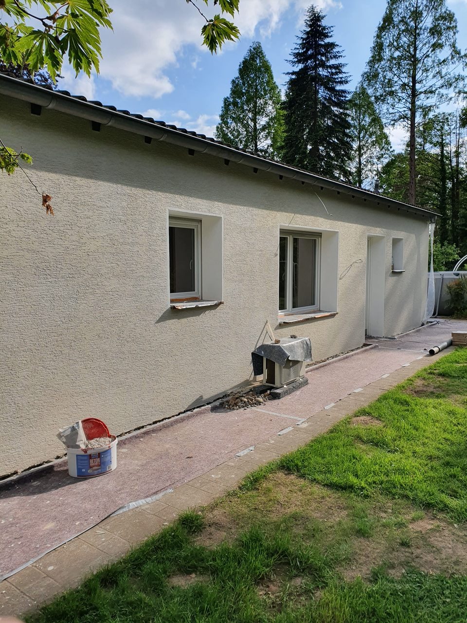 Elewacja budynku po nałożeniu tynku strukturalnego, widoczne okna i drzwi. Przy budynku stoi wiadro z zaprawą. Tło stanowią drzewa i błękitne niebo.