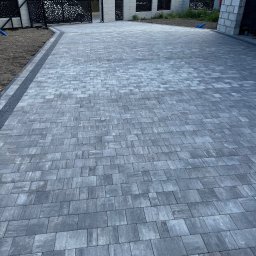 T&S Bruk Design s.c. - Ułożona szara kostka brukowa na podjeździe, widok z góry, z dekoracyjnym ogrodzeniem i wjazdem w tle. Nowoczesne wzornictwo.