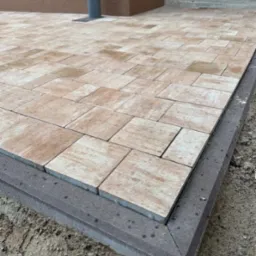 Beżowa kostka brukowa ułożona na posesji, obrzeża wykończone szarą kostką. Widok z góry, z bliska na fragment nawierzchni przy budynku i schodach.