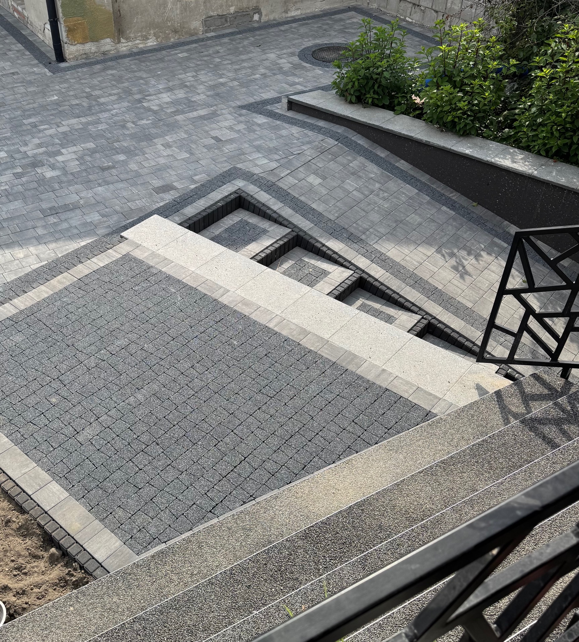 Ułożona kostka brukowa w odcieniach szarości, tworząca wzór schodów i placu. Widoczne elementy dekoracyjne z drobnego kamienia i roślinność w tle. Kąt ujęcia z góry.